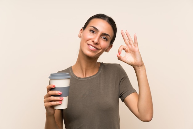 Die junge Frau, die ein hält, nehmen den Kaffee weg, der okayzeichen mit den Fingern zeigt