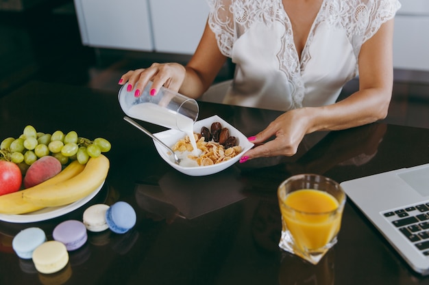 Die junge Frau gießt Milch in eine Schüssel mit Müsli zum Frühstück mit einem Laptop auf dem Tisch