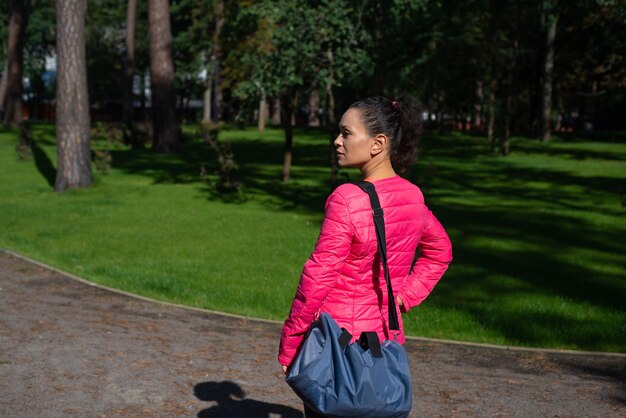 Die junge Frau mit einer Sporttasche auf der Schulter steht im Park. Seitenansicht.