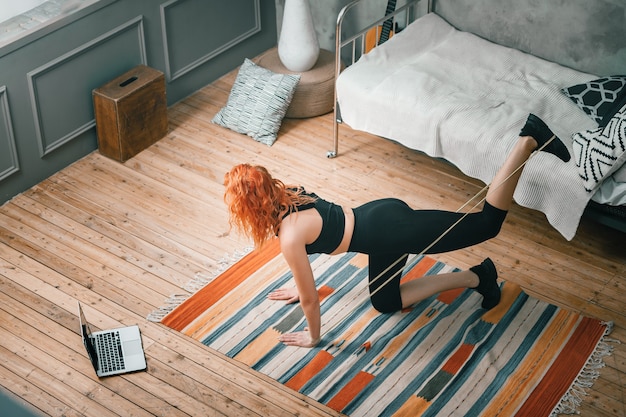 Die junge Frau treibt zu Hause Sport. Fröhliche Sportlerin mit roten Haaren macht Beine mit Sport-Fitness-Gummibändern, schaut sich einen Film an und lernt von einem Laptop im Schlafzimmer