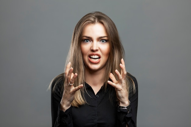 Die junge Frau wird wütend und schreit. Schöne Blondine in einem schwarzen Hemd. Negativität und Aggression. Grauer Hintergrund.