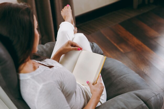 Die junge Frau zu Hause sitzt auf einem modernen Stuhl vor dem Fenster, entspannt sich in ihrem Wohnzimmer und liest ein Buch