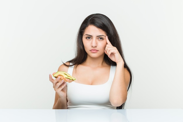 Die junge hispanische Frau, die einen Avocadotoast zeigt seinen Tempel mit dem Finger hält und denkt, konzentrierte sich auf eine Aufgabe.