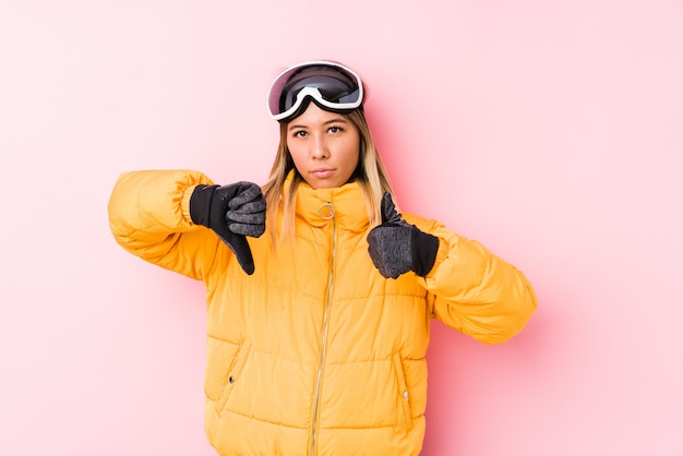 Die junge kaukasische Frau, die einen Ski trägt, kleidet in einer rosa Wand, die Daumen hoch und Daumen unten zeigt, wählen schwieriges Konzept