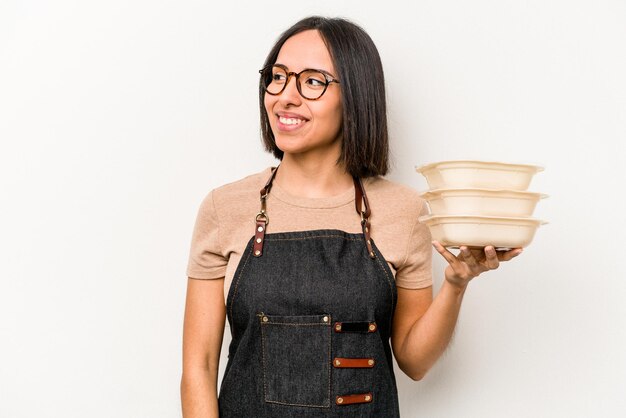 Die junge kaukasische Kellnerin, die Tupperware isoliert auf weißem Hintergrund hält, sieht fröhlich und angenehm lächelnd beiseite