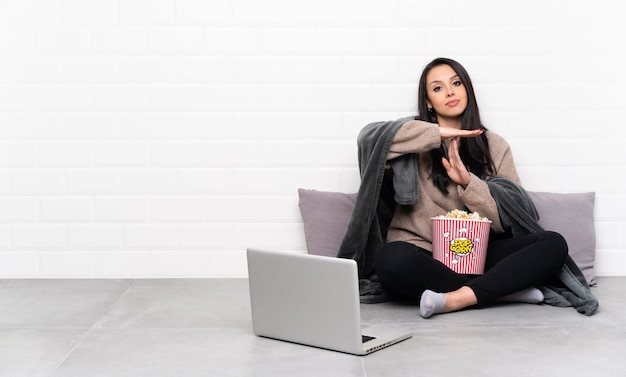 Die junge kolumbianische Frau, die eine Schüssel Popcorn hält und einen Film in einem Laptop macht Zeit heraus zeigt, gestikulieren