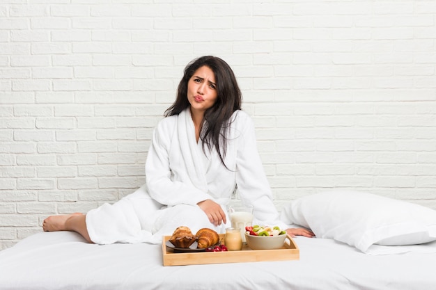 Die junge kurvige Frau, die ein Frühstück auf dem Bett nimmt, brennt Backen durch, hat müden Ausdruck. Gesichtsausdruck Konzept.