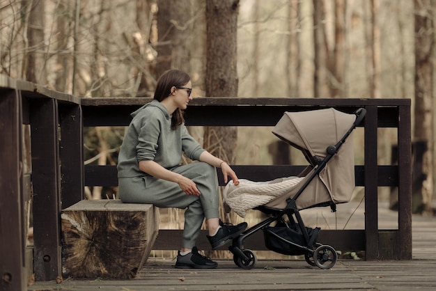 Die junge Mutter entspannt sich auf der Holzbank, während ihr Kleinkind im Kinderwagen schläft Die Mutter mit ihrem Kind im Park an der frischen Luft