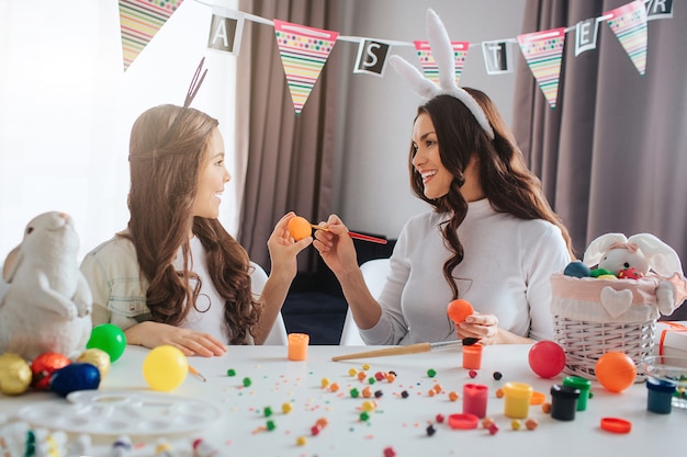 Die junge Mutter und ihre Tochter bereiten sich auf Ostern vor. Sie brechen sich gegenseitig Eier und lächeln. Junge Frau und ihre Tochter tragen Hasenohren. Dekoration und Malerei auf dem Tisch.