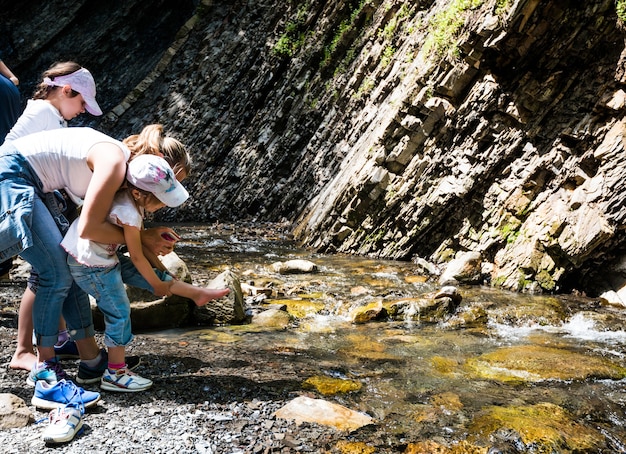 Die junge Mutter versucht ihrer Tochter zu helfen, ihre Schuhe auszuziehen, um sich beim Trekking im Gebirgsbach die Füße nass zu machen