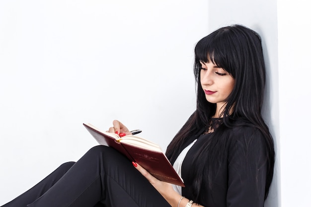 Die junge schöne ernste Brunettefrau, die auf einem Boden in einem Büro sitzt, schreibt in ein Notizbuch