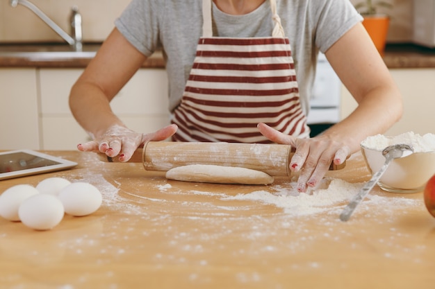 Die junge schöne glückliche frau, die an einem tisch mit mehl und tablette sitzt, einen teig mit einem nudelholz rollt und in der küche einen kuchen zubereitet. Kochen nach Hause. Essen aus nächster Nähe zubereiten.