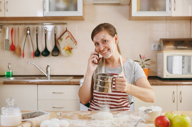 Die junge schöne glückliche Frau siebt Mehl mit einem Sieb, telefoniert mit dem Handy und sucht in der Küche nach einem Rezept für Kuchen in Tablette. Kochen nach Hause. Essen zubereiten.