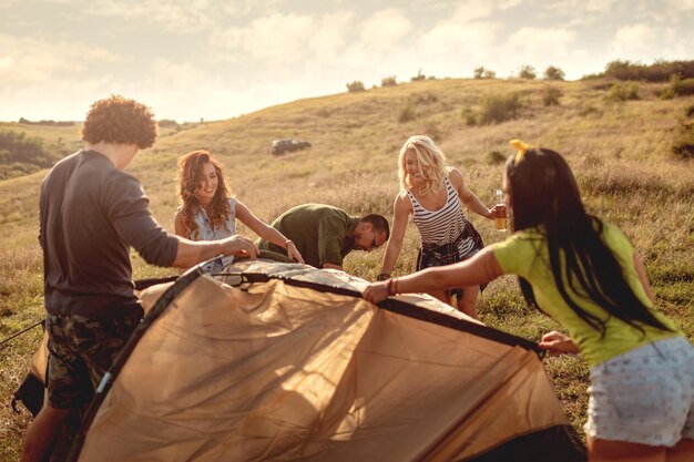 Die jungen glücklichen Freunde bereiten sich auf das Camping vor. An einem geeigneten Platz auf einer Wiese bauen sie ein Zelt auf und ihre Freundinnen helfen ihnen dabei.