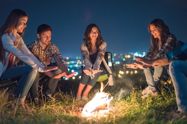 Die jungen Leute, die sich in der Nähe eines Lagerfeuers auf einem Hintergrund der Stadtbeleuchtung erwärmen. Nachtzeit