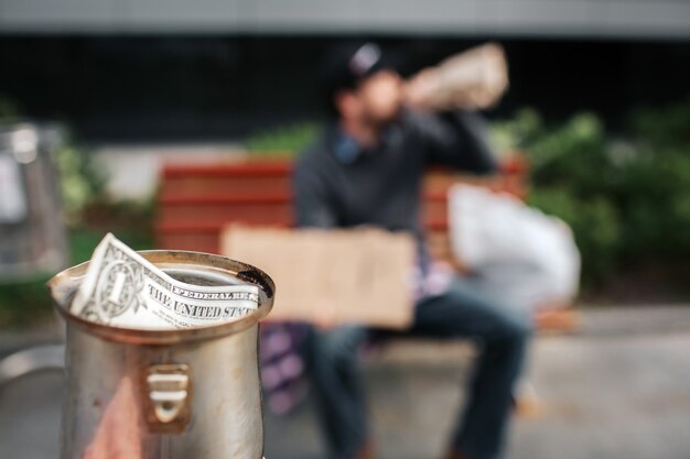 Die Kamera ist auf Metallbecher fokussiert. Da ist ein Dollarschein drin. Guy sitzt auf der Bank und trinkt aus der Flasche. Es ist seine Tasse.