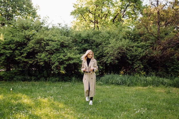 Die kaukasische blonde frau trägt ein grabenlächeln glücklich am sonnigen frühlingstag draußen, der im park geht