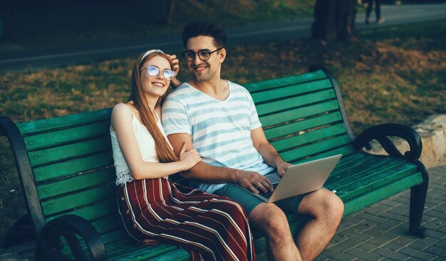 Die kaukasische rothaarige Freundin mit Sommersprossen und Brille sitzt in der Nähe ihres Geliebten auf der Bank, während sie einen Laptop benutzt