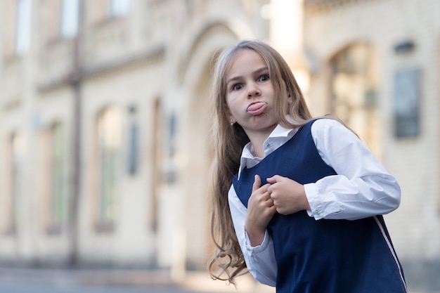 Die Kindheit ist voller Lernen. Lustiges Kind streckt die Zunge heraus. Kleine Mädchen tragen Uniform im Freien. Zurück zur Schule. Kinderbetreuung und Bildung. Kindertagesstätte. Internationaler Kindertag, Kopierraum.