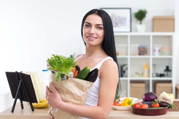 Die Köchin, die eine Tasche voller Grüns hält, kam gerade vom Markt zurück, bereit, gesundes Essen mit einem Kochbuch-Rezept, Nahaufnahme, Vorderansicht, Porträt zuzubereiten