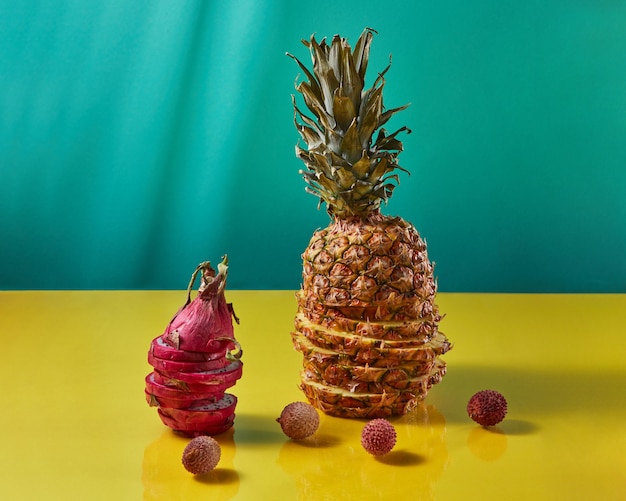 Die Komposition mit tropisch-exotischen Früchten einzelner Ananas und Drachenfrucht, Pitaya aus Scheiben und Litschifrüchten auf duotongelb-blauem Hintergrund.