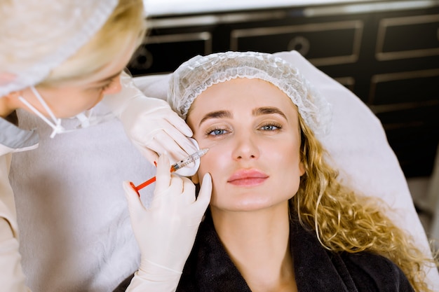 Die Kosmetikerin des Arztes führt das verjüngende Verfahren für Gesichtsspritzen durch, um Falten auf der Gesichtshaut einer schönen jungen Frau in einem Schönheitssalon zu straffen und zu glätten.