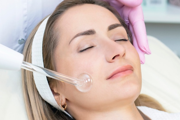 Die Kosmetikerin führt ein Pulsstromverfahren für das Gesicht einer jungen Frau durch. schließen