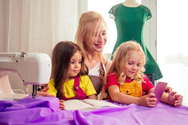 Die lächelnde, glückliche, Multitasking-blonde Mutter bringt einer kleinen Schwester bei, selbst an einer Nähmaschine zu nähen und Selfie-Fotos auf dem Smartphone zu machen