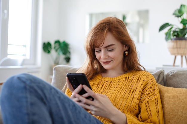 Die lächelnde hübsche junge Frau entspannt sich auf dem Sofa in ihrem Wohnzimmer, während sie ihr mobiles Smartphone für soziale Medien nutzt und im Internet surft