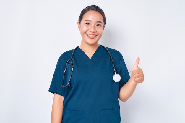 Die lächelnde junge asiatische Krankenschwester, die blaue Uniform mit einem Stethoskop trägt, hat alles unter Kontrolle und zeigt Thumbsup in Zustimmung, die auf weißem Hintergrund isoliert ist. Konzept der Gesundheitsmedizin