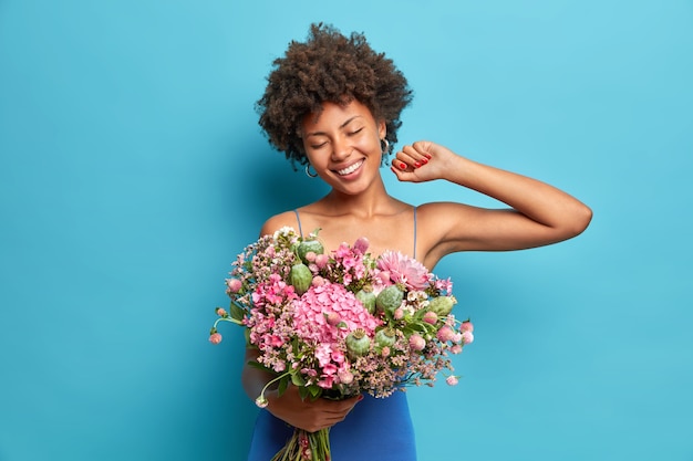 Die lächelnde sorglose Afroamerikanerin mit den lockigen Haaren feiert die Posen des 8. März mit einem schönen Blumenstrauß und trägt ein festliches Kleid, das über der blauen Wand isoliert ist