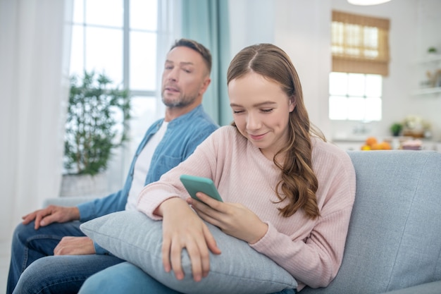 Die lächelnde Tochter mit dem Smartphone in den Händen und dem Blick auf den Bildschirm wandte sich von ihrem Vater ab, die beide auf der Couch saßen.