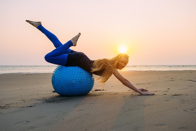 Die langhaarige Blondine ist während des Sonnenuntergangs mit Pilates auf einem Trainingsball am Strand beschäftigt