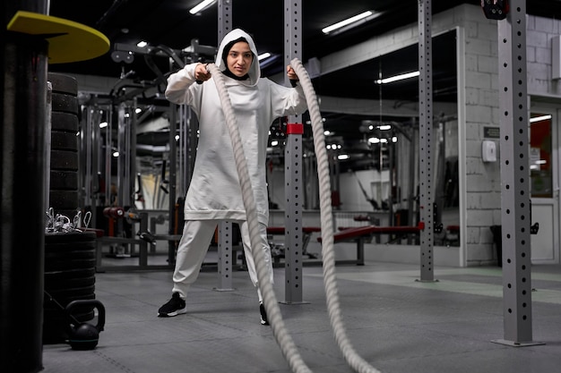 Die leistungsstarke arabische CrossFit-Trainerin im Hijab trainiert allein im Fitnessstudio mit Seilen und konzentriert sich auf Übungen mit sportlichen Geräten