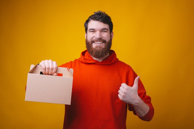 Die Lieferung von Essen ist super. Bärtiger Mann hält eine Box mit Platz für Ihren Text und zeigt Daumen nach oben. Bleib zu Hause und bestelle.