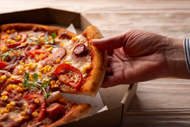 Die menschliche kaukasische Hand nimmt ein Stück große Pizza aus einem Karton auf einem weißen Holztisch