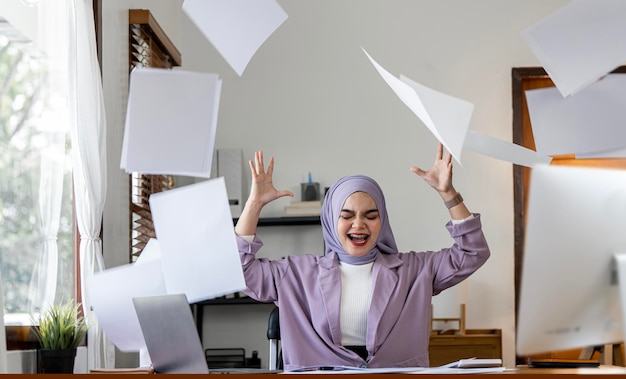 Die muslimische Millennial-Büroangestellte schreit, schreit und wirft Papierkram an ihrem Schreibtisch hoch Depression überarbeitetes Versagen