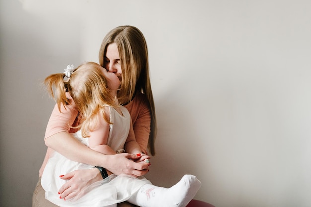 Die Mutter küsst kleines Mädchen auf hellem Hintergrund im Innenbereich Porträt von Mutter und Tochter, die zu Hause sitzen Das Konzept des glücklichen Familienurlaubs Frauentag Muttertag