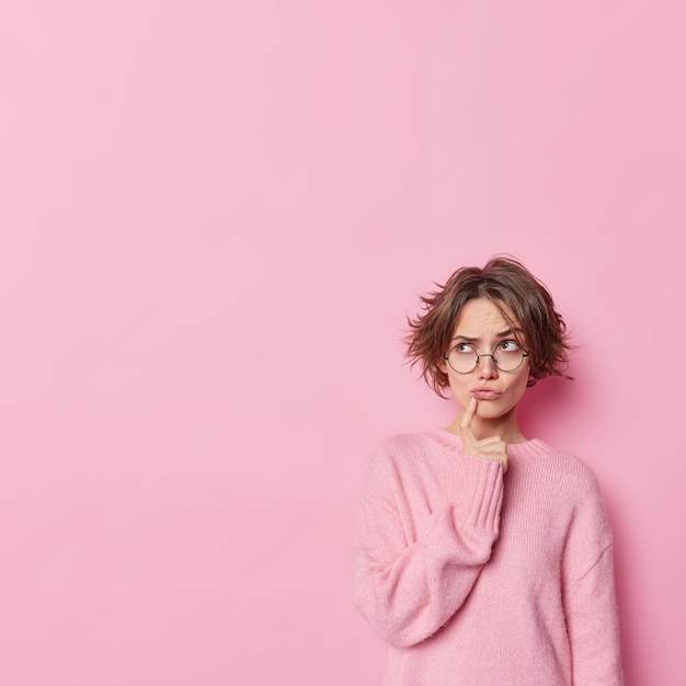 Die nachdenkliche schöne junge Frau hält den Zeigefinger in der Nähe der Lippen und denkt über etwas nach, das eine runde Brille trägt, und ein Pullover steht vor einem rosafarbenen Studiohintergrund mit Leerzeichen für Ihre Werbung