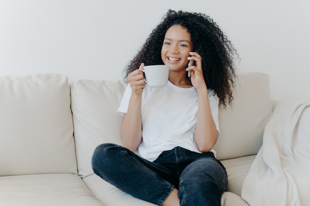 Die nette afroamerikanische Frau, die Kaffeepause im Wohnzimmer hat, sitzt auf Couchanrufen Freund über modernes Gerät