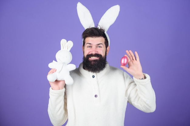 Die Osterhasenjagd beginnt. Glücklicher Mann mit Hasenohren, der Hasenspielzeug und Ei hält. Bärtiger Mann im Kaninchenkostüm mit Osterei und Hasenspielzeug. Frühling, neues Leben und Fruchtbarkeit. Frühlingsferienfeier.