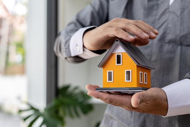 Die Person legt ihre Hand auf ein Miniaturhausmodell, ein Foto des Hausversicherungskonzepts, beim Kauf eines neuen Hauses sollte eine Hausratversicherung abgeschlossen werden, um sicher zu sein, dass eine Gefahr für das Haus besteht.