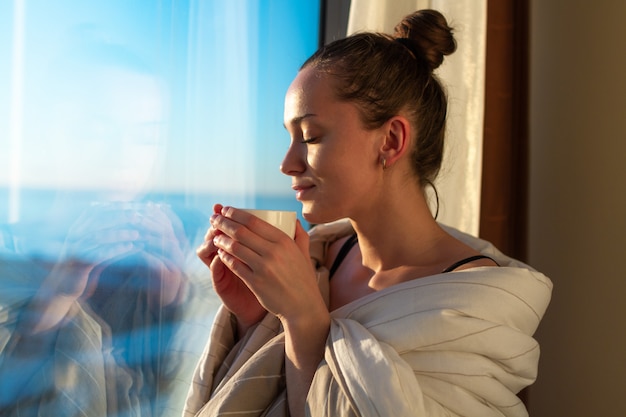 Die recht junge Frau, die in eine Decke eingewickelt wird, steht nahes Fenster und genießt Kaffee des ersten Morgens auf Sonnenschein. Ein frühes Aufwachen und der Beginn eines neuen Tages