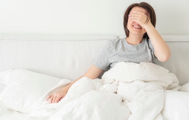 Die recht junge lächelnde Frau, die ihr Gesicht mit bedeckt, hatte das Sitzen auf dem Bett. Glückliche Dame, die Morgen und das Lachen genießt