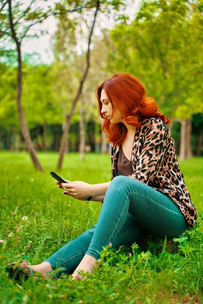 Die rothaarige Frau schreibt eine SMS, während sie auf dem niedlichen Mädchen des Grases sitzt und Fotos von grünem Gras im Park macht