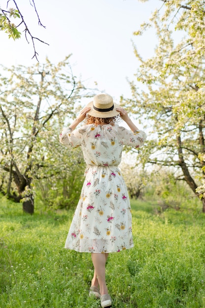 Die Rückansicht einer jungen Frau mit lockigem Haar setzt einen stilvollen Weidenhut auf, während sie in einem grün blühenden Garten geht. Frühlingsstimmung