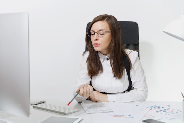 Die schöne ernsthafte und versunkene braunhaarige Geschäftsfrau in Anzug und Brille sitzt am Schreibtisch und arbeitet am Computer mit modernem Monitor mit Dokumenten im hellen Büro,