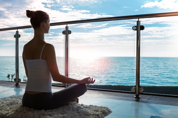 Die schöne Frau, die Yoga auf einer Gymnastikmatte praktiziert und Assana Yoga Pose auf einem Balkon mit Meerblick sitzt.