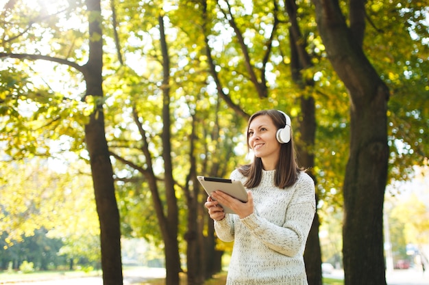 Die schöne fröhliche, fröhliche braunhaarige Frau im weißen Pullover mit einem Tablet, das an einem warmen Tag Musik in den weißen Kopfhörern im Herbstpark hört. Herbst in der Stadt.