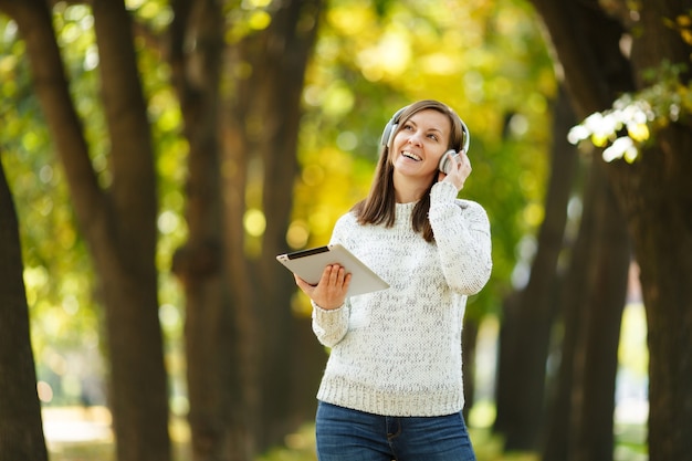 Die schöne fröhliche, fröhliche braunhaarige Frau im weißen Pullover mit einem Tablet, das an einem warmen Tag Musik in den weißen Kopfhörern im Herbstpark hört. Herbst in der Stadt.